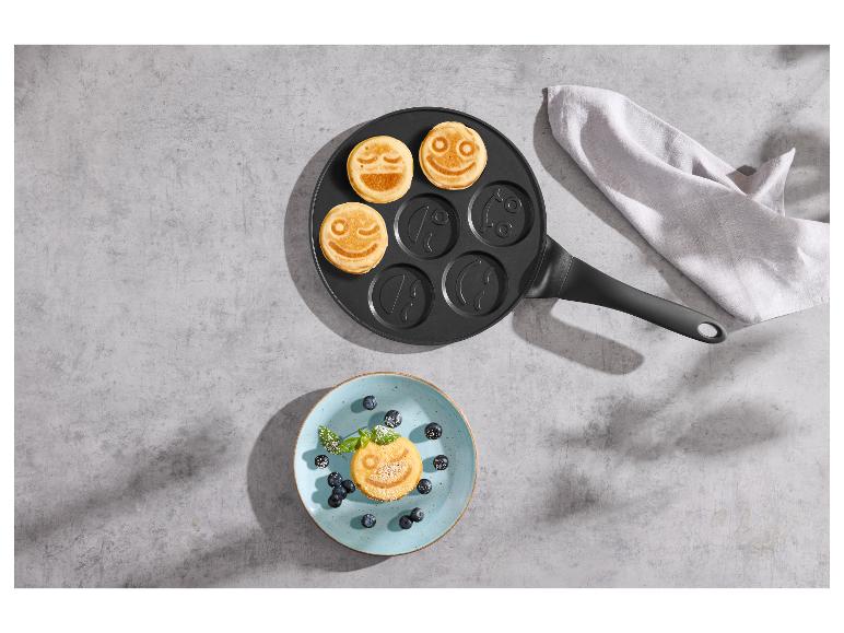 Pfannkuchenpfanne mit Smileys und Teller mit Pfannkuchen und Blaubeeren auf Betonhintergrund.