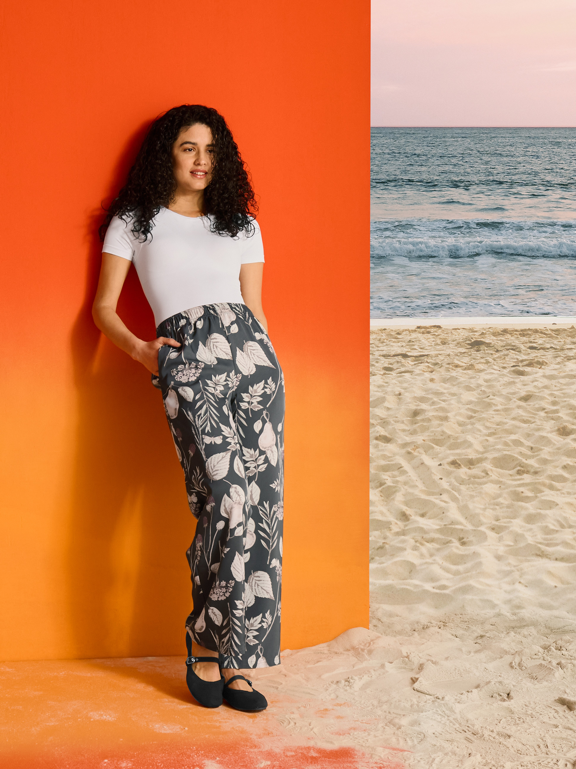 Frau in floraler Hose und weißem T-Shirt, vor orangefarbenem Hintergrund und Strand.