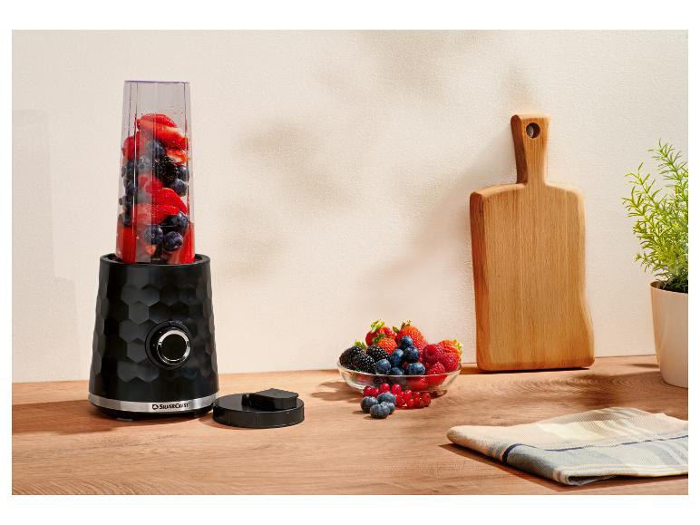 Schwarzer Mixer mit Früchten, eine Schale Beeren und ein Holzschneidebrett auf der Arbeitsplatte.