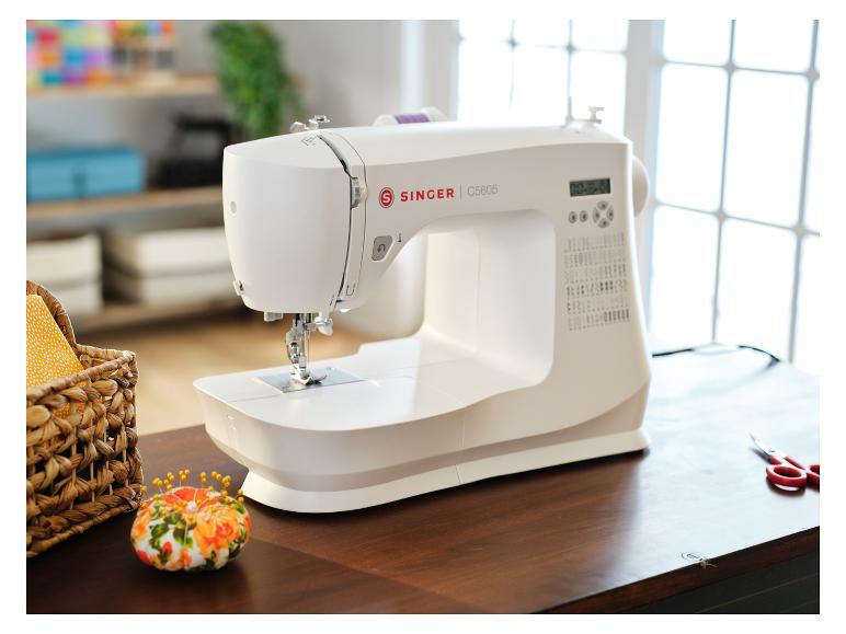 Weiße Singer-Nähmaschine auf einem Holztisch, mit einem Korb voller Zubehör und einem Nadelkissen.