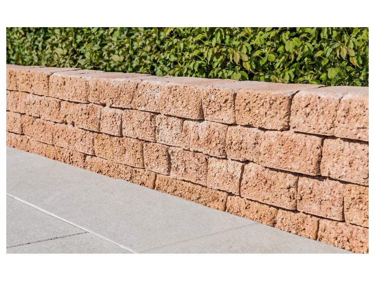 Stützmauer aus Betonblöcken mit einer grünen Hecke im Hintergrund und Terrassenplatten.