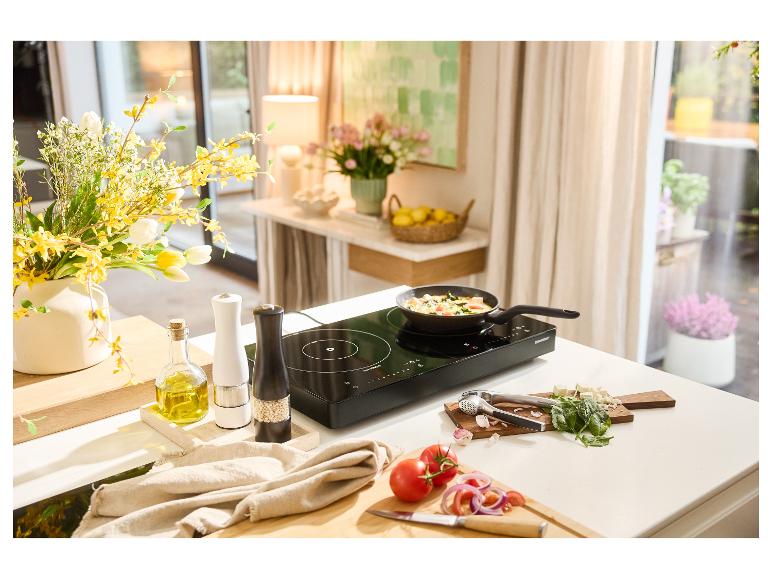 Ein schwarzes Induktionskochfeld mit einer Pfanne, die Gemüse kocht, umgeben von frischen Zutaten.