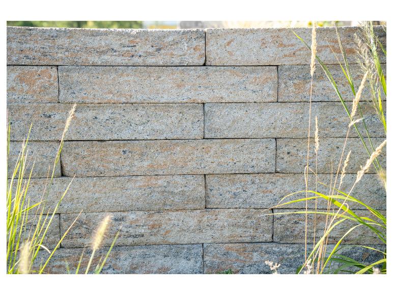 Nahaufnahme einer Steinmauer mit hellen rechteckigen Blöcken, teilweise von hohem Gras verdeckt.