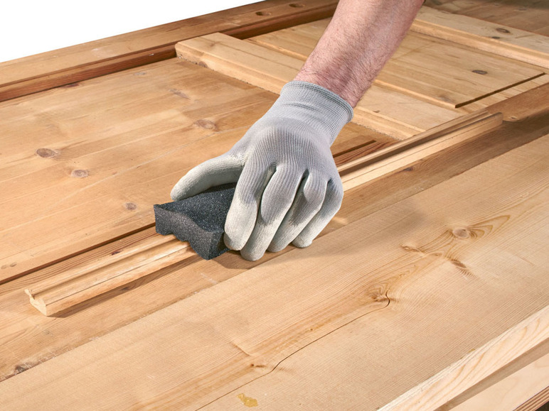 Eine Hand in einem Handschuh schleift ein Holzpaneel mit einem Schleifschwamm.