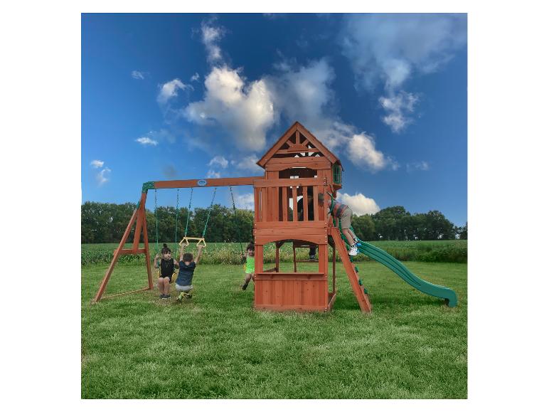 Kinder spielen auf einem Holzschaukelset mit Rutsche auf einem grünen Feld unter blauem Himmel.