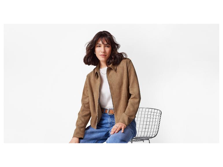 Eine Frau in einer braunen Wildlederjacke und Jeans sitzt auf einem Stuhl.