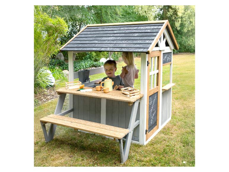 Zwei Kinder spielen in einem Holzspielhaus mit angebautem Picknicktisch und Küchenzubehör.