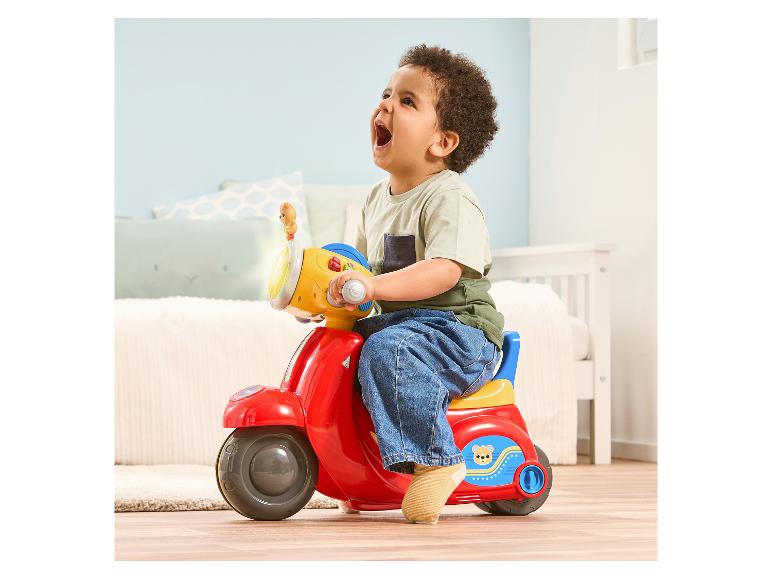 Lächelndes Kind sitzt auf einem roten Roller in einem Zimmer.