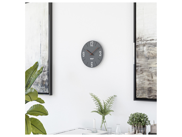 Eine graue Mebus Wanduhr hängt an einer weißen Wand.