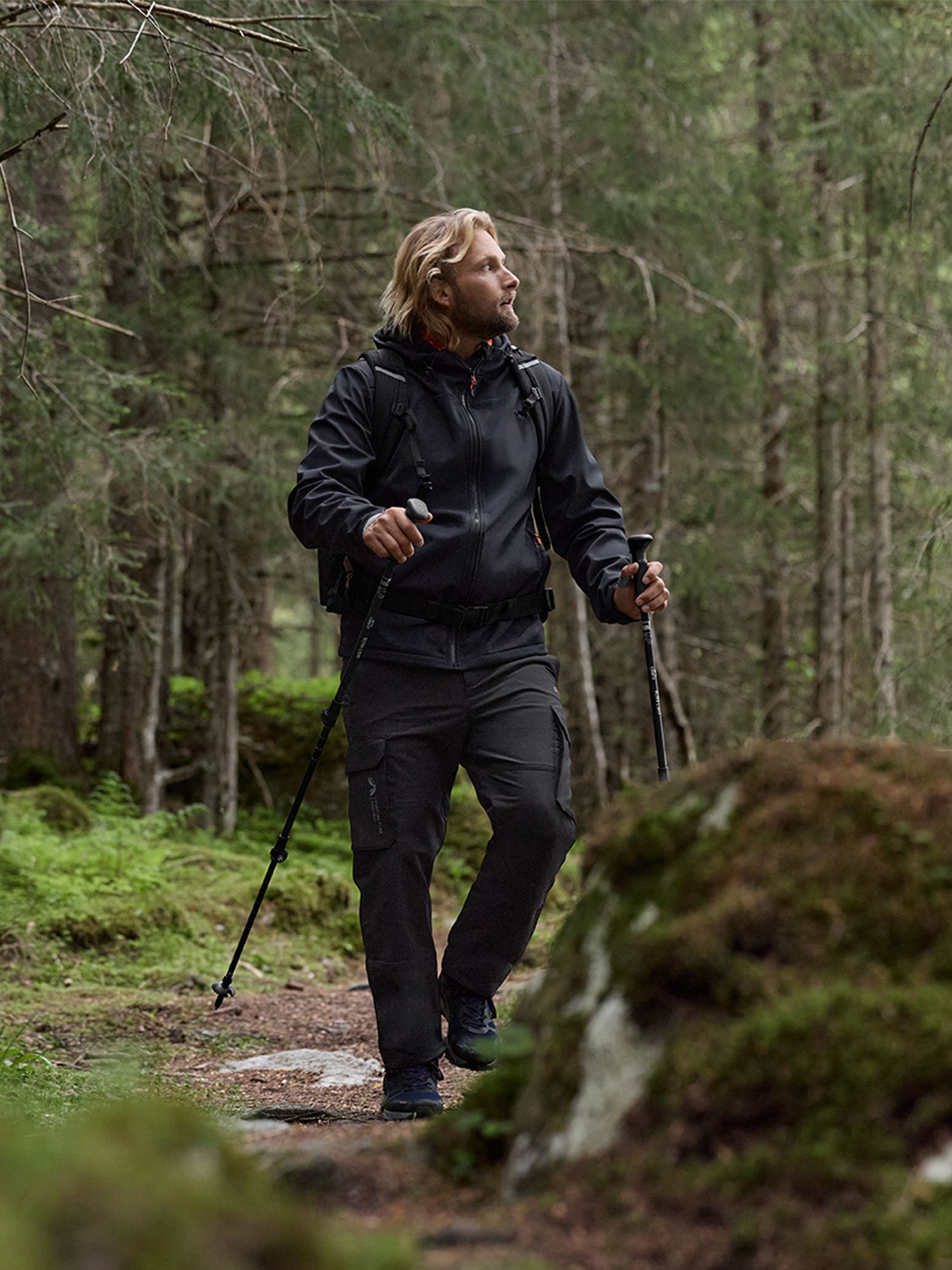 Ein Mann in schwarzer Wanderjacke und Cargohose geht mit Trekkingstöcken in einem Wald.
