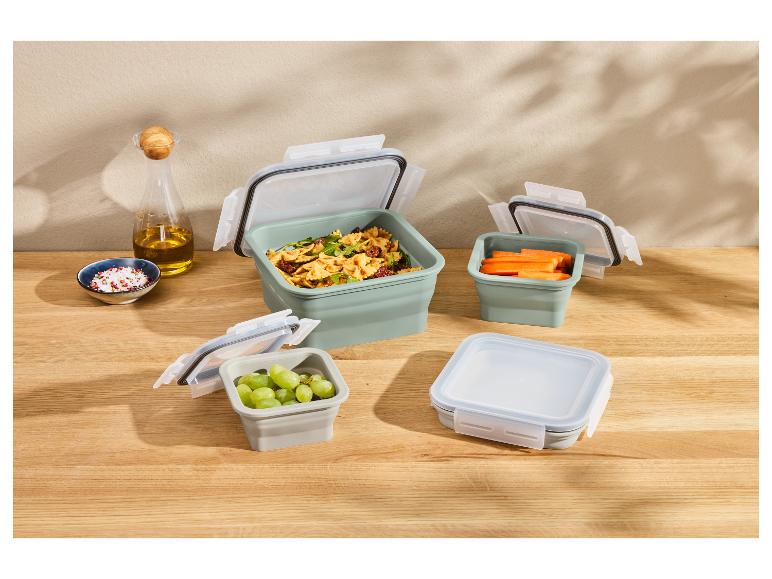 Faltbare Lebensmittelbehälter mit Nudeln, Trauben und Karotten auf einem Holztisch.