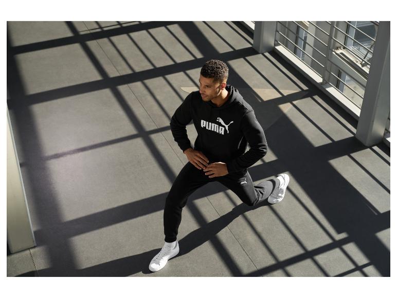 Mann in schwarzem Hoodie und Jogginghose mit weißem Logo, macht einen Ausfallschritt.
