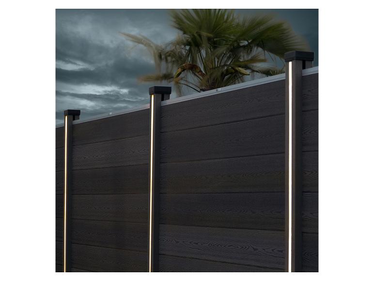 Dunkler Holzzaun mit vertikalen LED-Leuchten und einer Palme im Hintergrund.