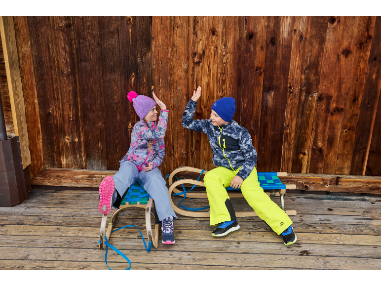 Zwei Kinder in Winterkleidung und Mützen, sitzen auf Schlitten und geben sich High-Five.