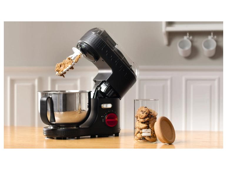 Eine schwarze Küchenmaschine mit Teig in der Schüssel und ein Glas Kekse auf einem Holztisch.