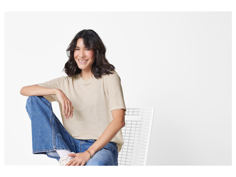 Eine lächelnde Frau in einem beigen Shirt und Jeans sitzt auf einem weißen Stuhl.