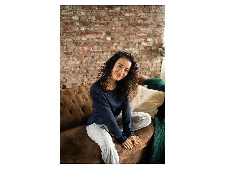 Frau in blauem Oberteil und Pyjamahose mit Weihnachtsmuster, sitzend auf einer braunen Couch.