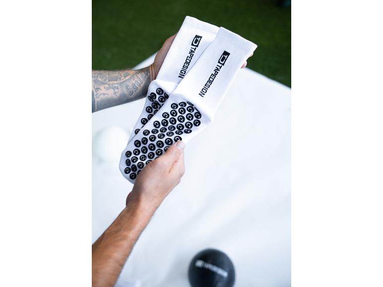 Hände halten weiße Sportsocken mit schwarzen Anti-Rutsch-Noppen und „TAPEDESIGN“-Logo.