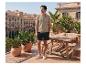 Mann in beigem Poloshirt und dunklen Shorts auf einer sonnigen Terrasse mit Stadtblick.