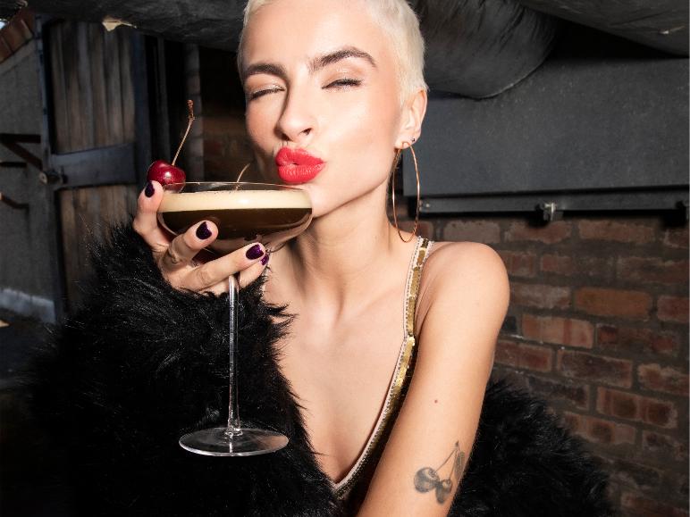 Frau mit kurzen Haaren und rotem Lippenstift hält einen Cocktail mit Kirsche.