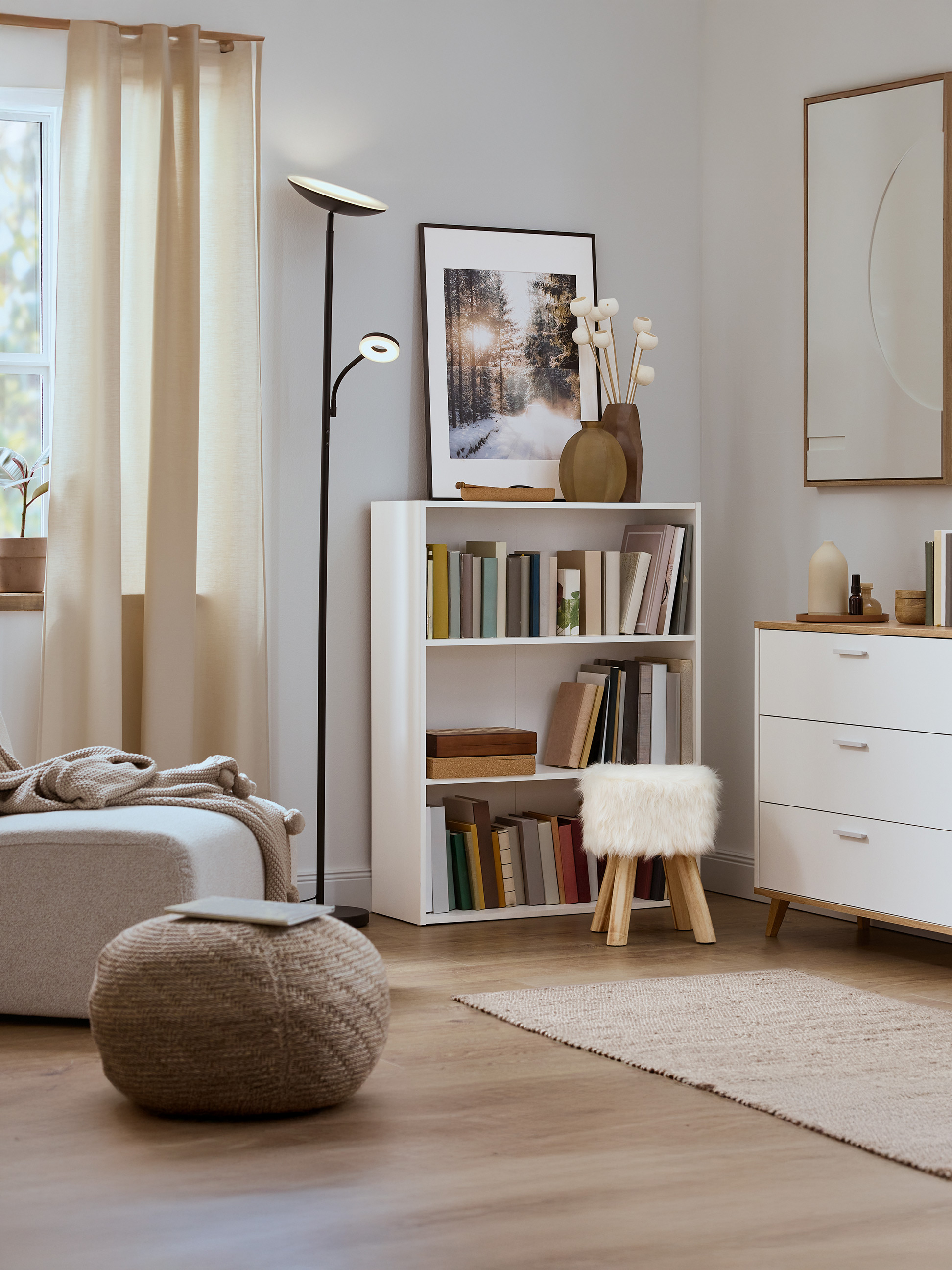 Helles Wohnzimmer mit weißem Bücherregal, Strickpouf und moderner Stehlampe.