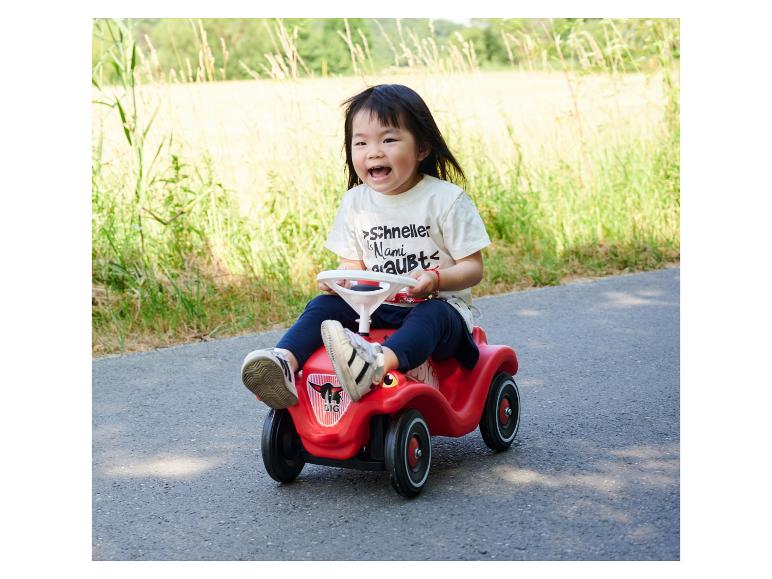 Lachendes Kleinkind in einem roten Spielzeugauto auf einem Weg mit Gras im Hintergrund.