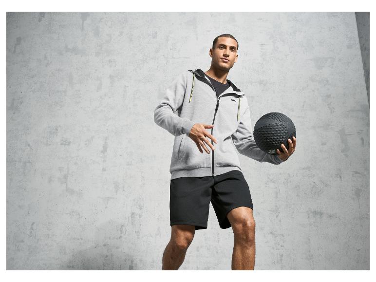 Mann in grauem Hoodie und schwarzen Shorts, hält einen schwarzen Medizinball fest.