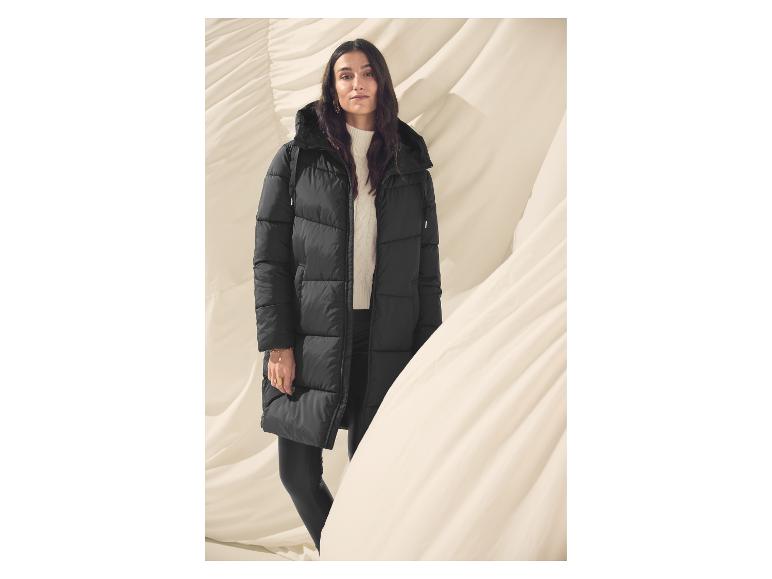 Eine Frau in schwarzem Steppmantel und weißem Pullover steht vor einem drapierten Stoffhintergrund.