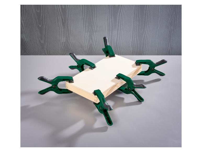 Sechs grüne Federklemmen halten einen Holzrahmen auf grauem Hintergrund.