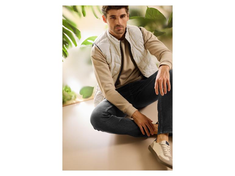 Mann in beiger Steppweste, Sweatshirt und dunklen Jeans, sitzend in natürlicher Umgebung.