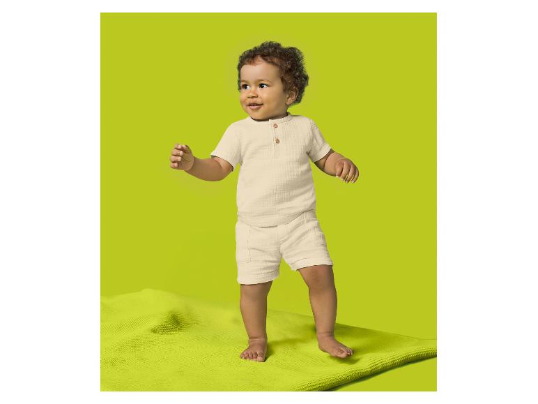 Kleinkind in cremefarbenem Kurzarmhemd und Shorts, stehend auf einer grünen Matte.