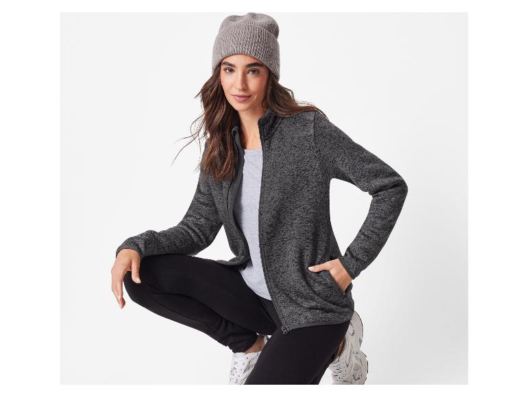 Frau in grauer Jacke, schwarzer Hose und grauer Mütze, hockend.