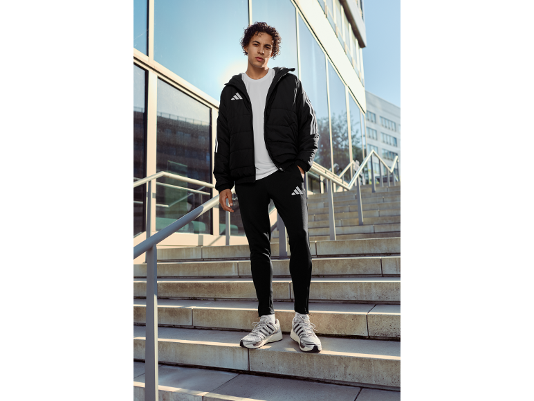 Mann in schwarzer Steppjacke, weißem T-Shirt, schwarzer Jogginghose und Turnschuhen.