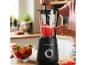 Frau bereitet Erdbeer-Smoothie in schwarzem Mixer zu, umgeben von frischen Erdbeeren.