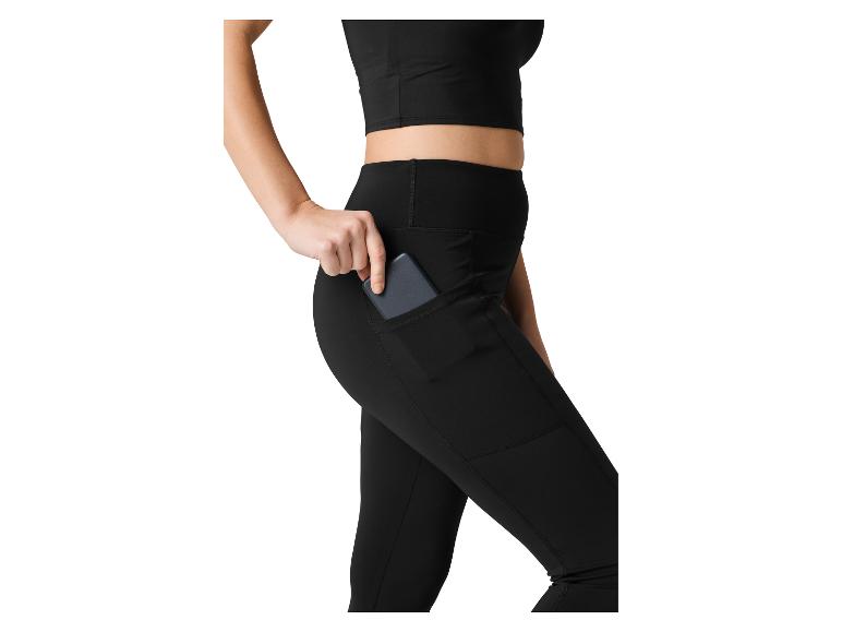 Frau in schwarzem Crop-Top und Leggings, steckt ein Telefon in die Seitentasche.