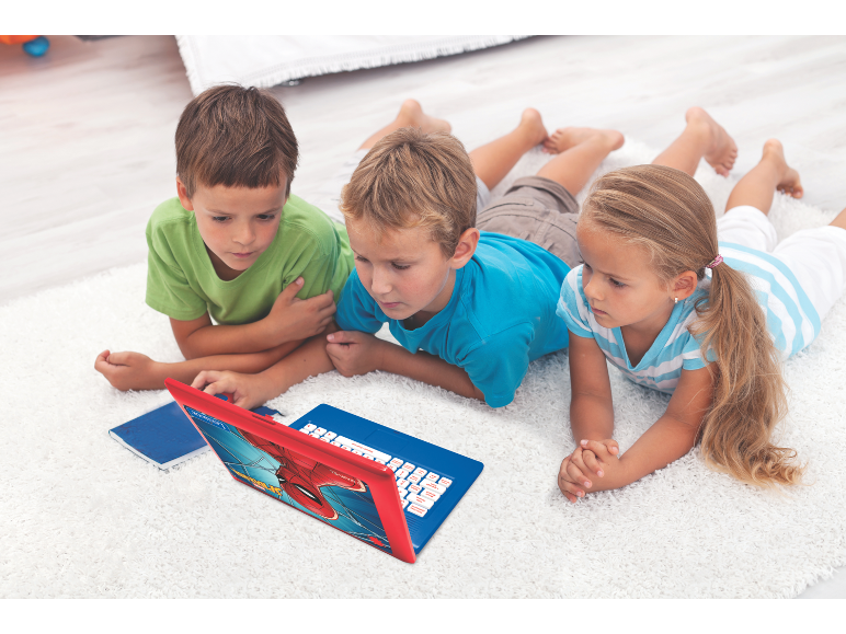 Drei Kinder liegen auf einem weißen Teppich und schauen auf einen rot-blauen Spielzeuglaptop.