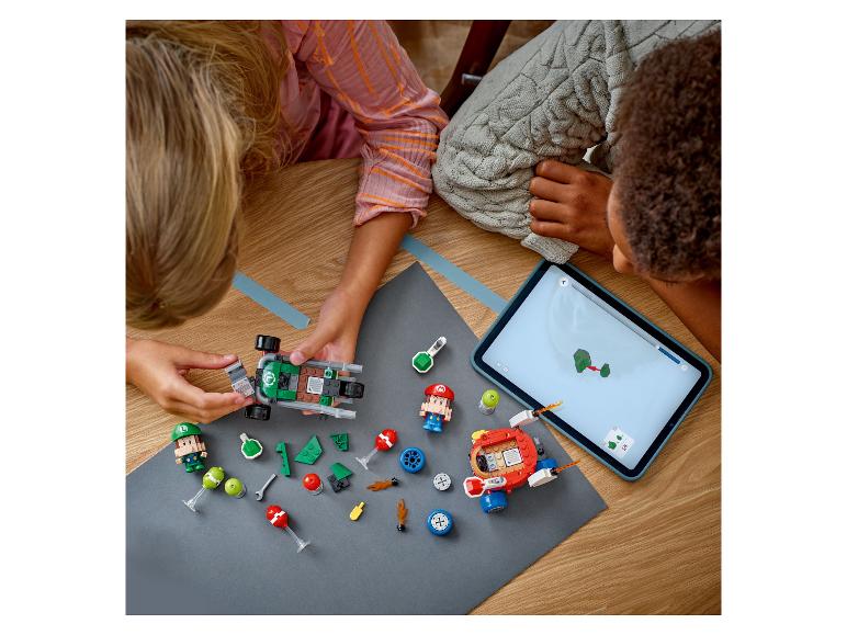 Zwei Kinder spielen mit LEGO Super Mario Sets, darunter ein Kart und Figuren, angeleitet von einem Tablet.