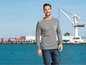 Mann in einem grauen Pullover und blauen Arbeitshose vor einem Hafen.