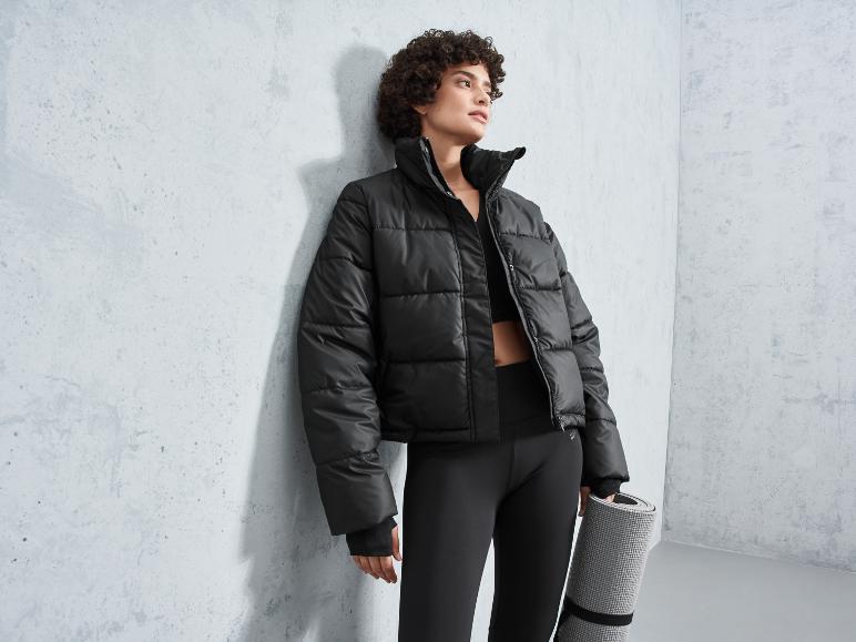 Frau in einer schwarzen Daunenjacke und Leggings hält eine Yogamatte.
