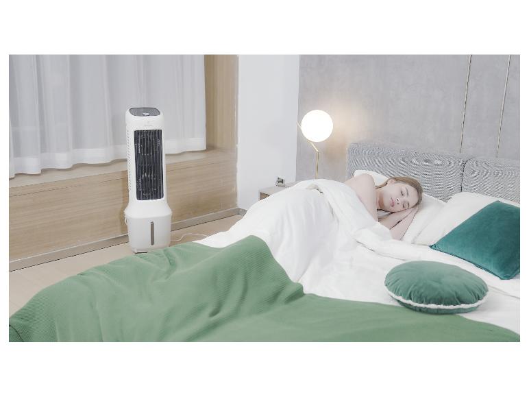 Ein weißer Turmventilator neben einem Bett mit einer Frau, die unter einer grünen Decke schläft.