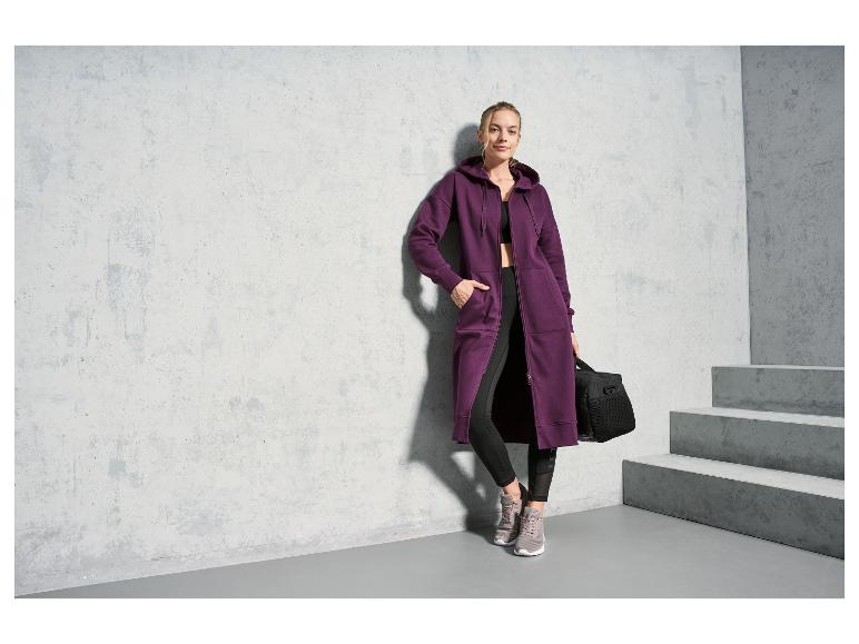 Eine Frau in einem lila Hoodie und schwarzen Leggings mit einer Sporttasche.