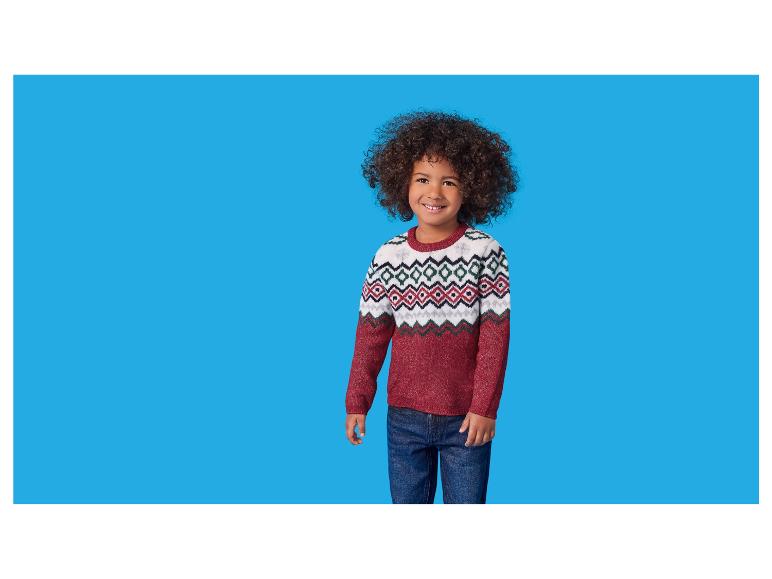 Ein lächelnder Junge mit lockigem Haar, der einen rot-weiß gemusterten Pullover und Jeans trägt.