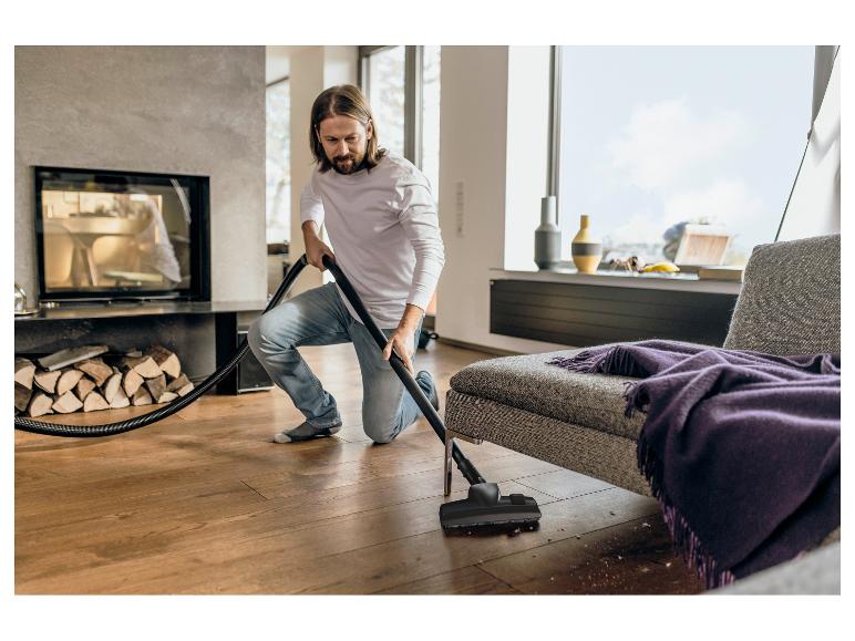 Mann saugt Holzboden, mit Kamin und Sofa im Hintergrund.