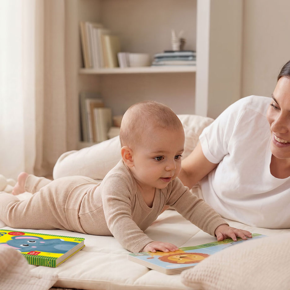 Baby im beigefarbenen Body spielt mit bunten Tierbüchern auf einer Matte, mit lächelndem Erwachsenen.
