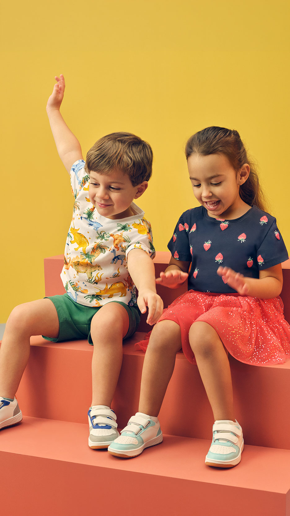 Zwei Kinder sitzen auf Stufen, Junge in Dinosaurier-T-Shirt, Mädchen in Erdbeer-T-Shirt.