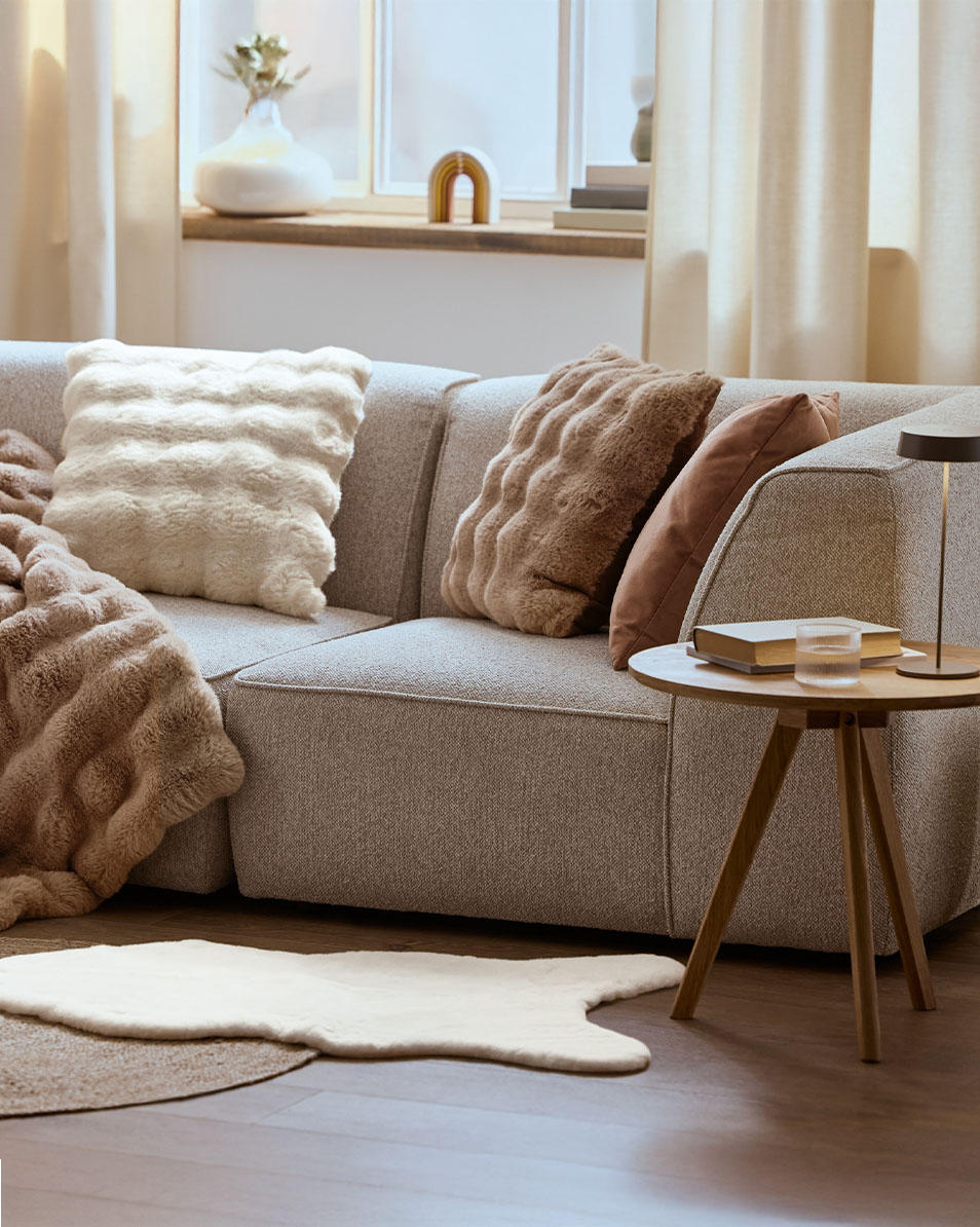 Gemütliches Wohnzimmer mit grauem Sofa, weichen Kissen und Decken in neutralen Farben.