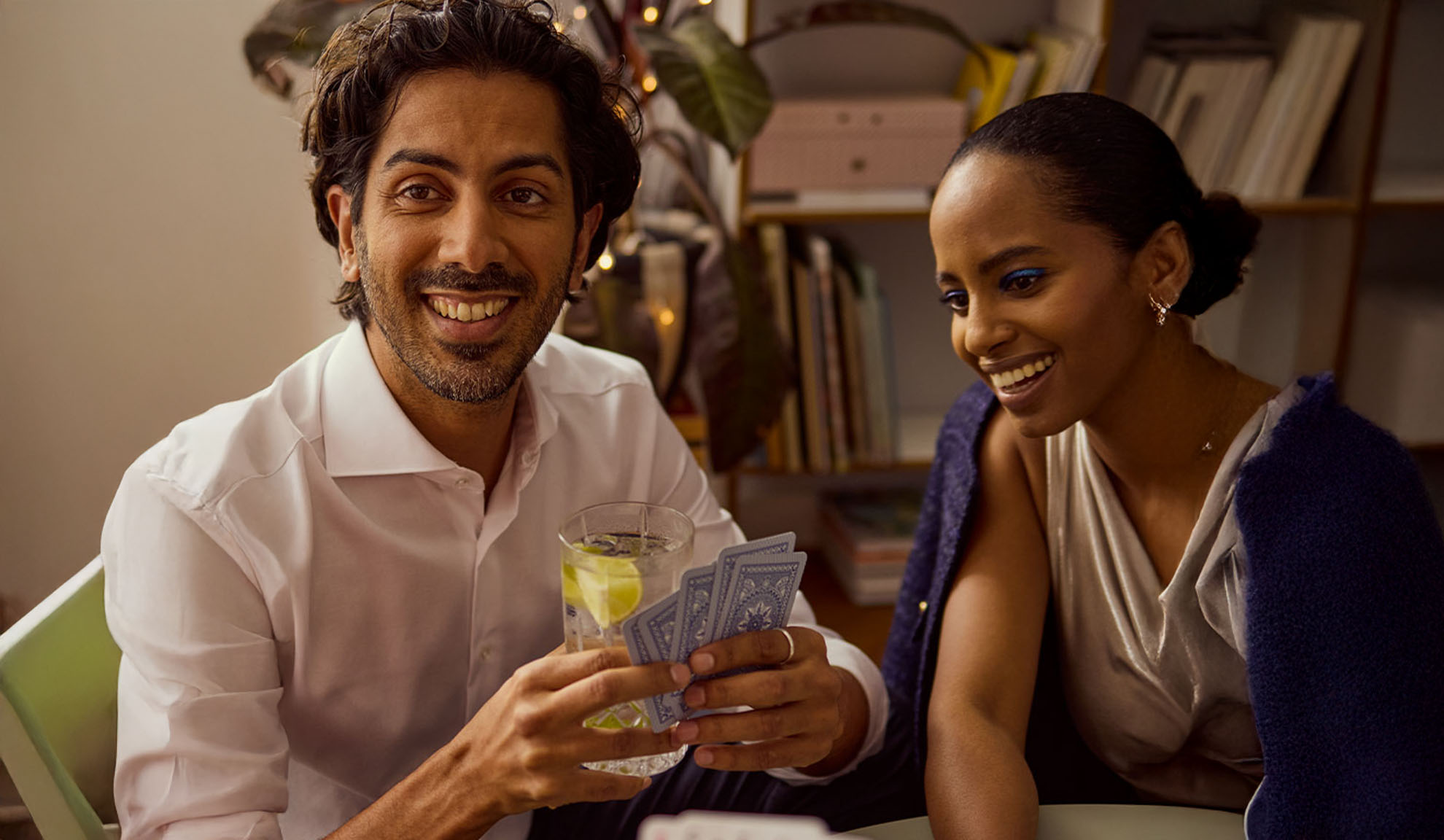 Mann und Frau spielen Karten, der Mann hält ein Getränk und Karten.