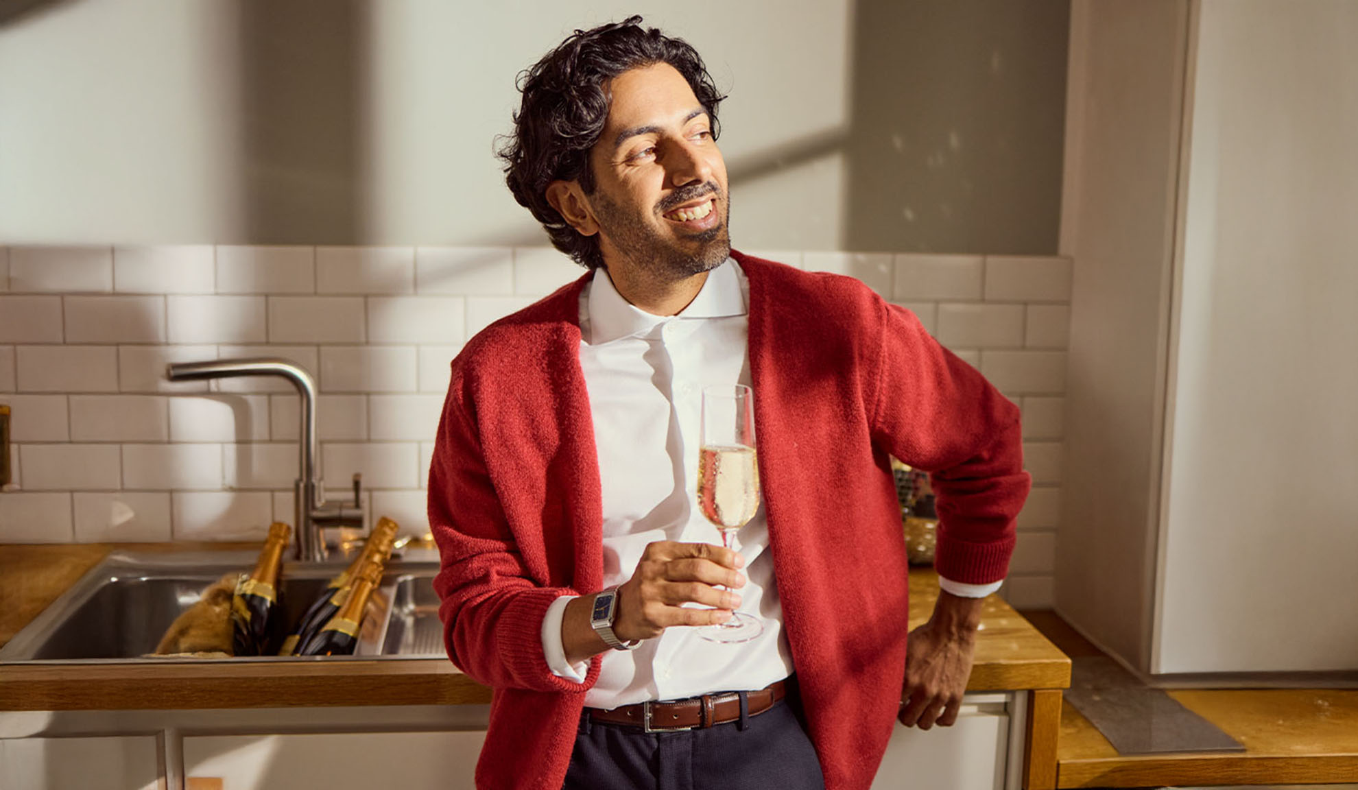 Lächelnder Mann in roter Strickjacke, der ein Glas Sekt in einer Küche hält.