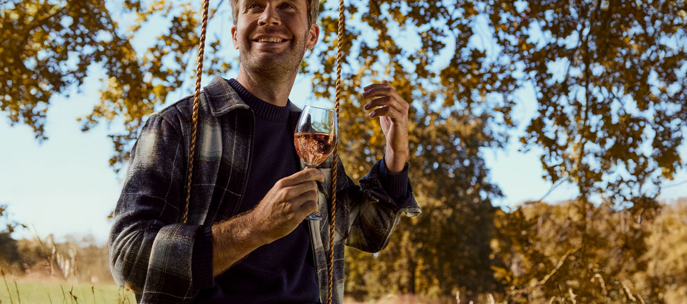 Mann in kariertem Hemd und Pullover hält ein Glas Roséwein auf einer Schaukel im Freien.