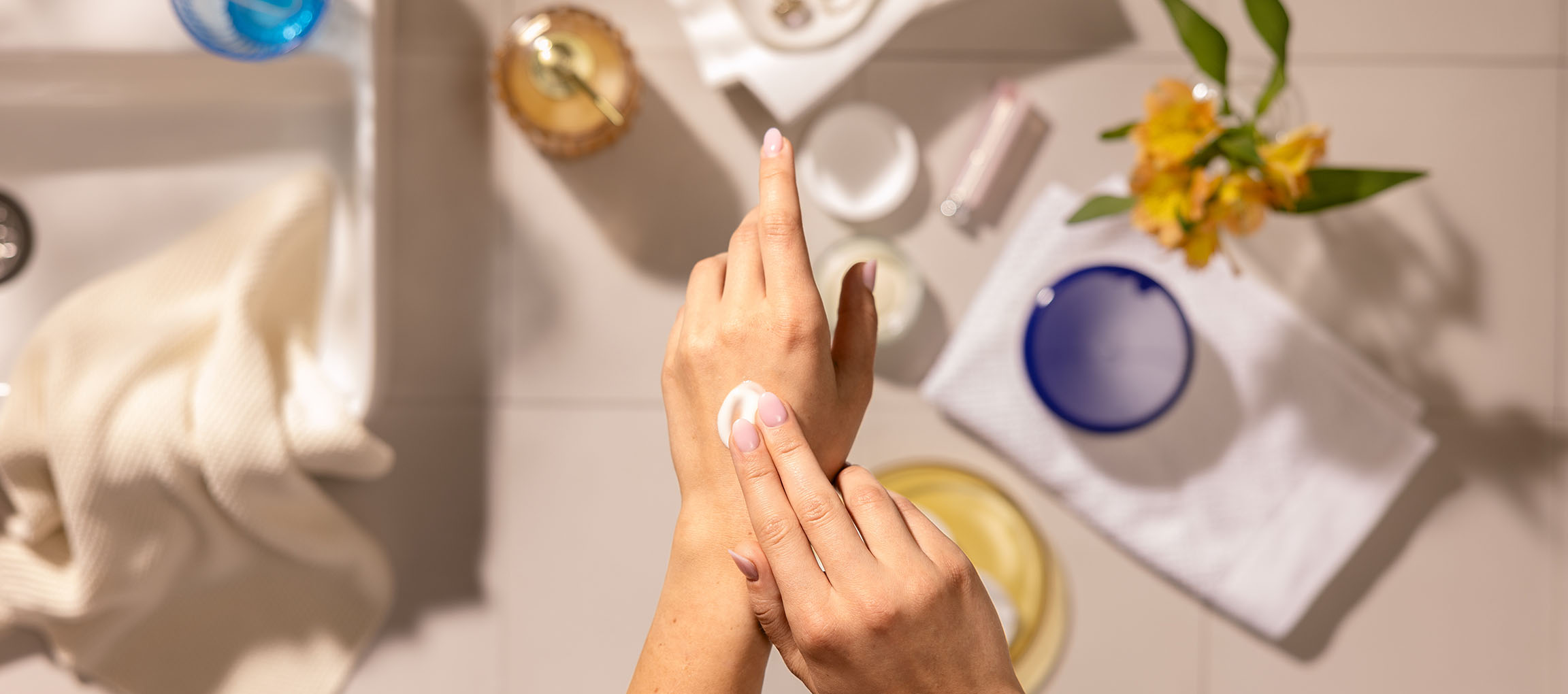 Frauenhände tragen Handcreme auf, umgeben von Kosmetik und Handtüchern im Badezimmer.